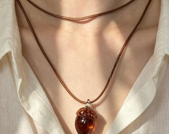 Puede incluir: Un colgante con forma de bellota marrón hecho de material translúcido color ámbar con una tapa metálica detallada. El colgante cuelga de un collar de cordón marrón de doble capa, sobre una camisa blanca de cuello con textura visible.