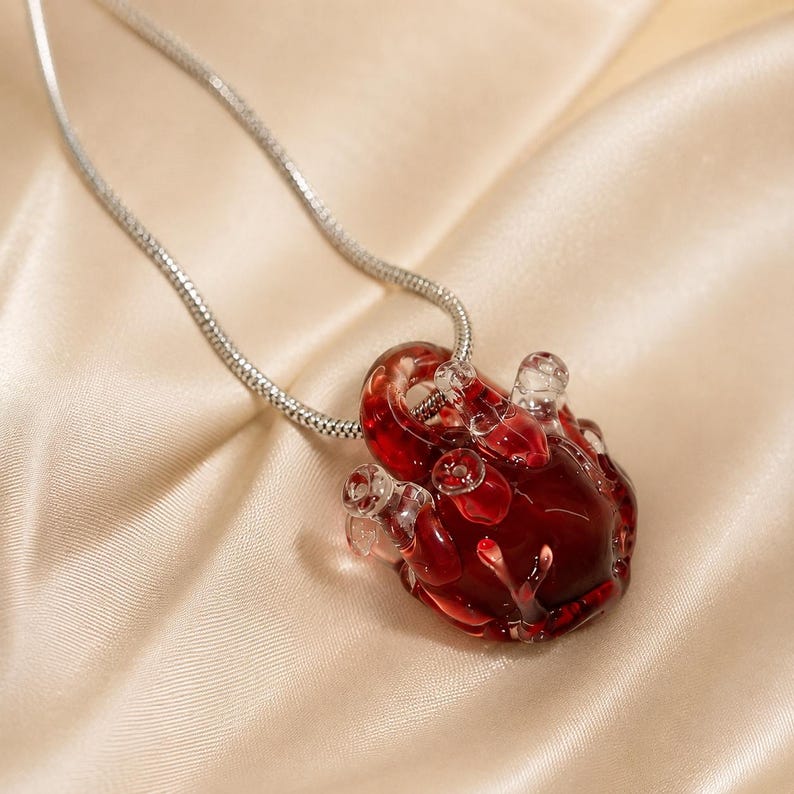 Puede incluir: Un collar con un colgante de vidrio transl&uacute;cido de color rojo intenso con forma de coraz&oacute;n anat&oacute;mico y detalles en relieve que recuerdan a vasos sangu&iacute;neos. El colgante cuelga de una fina cadena de serpiente de metal plateado flexible.