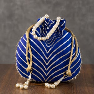 Könnte beinhalten: Ein königsblauer Beutel mit Kordelzug und einem Chevron-Muster aus silbernen Metallic-Linien. Die Tasche verfügt über einen Griff aus weißen Kunstperlen und zwei goldfarbene, geflochtene Kordeln mit hängenden weißen Perlenquasten.