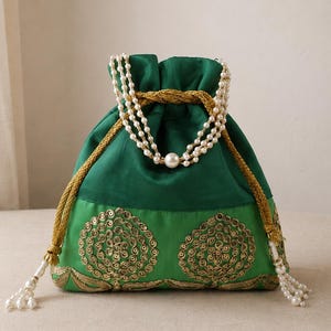 Può includere: Una borsa a sacchetto con coulisse verde dal design in tessuto bicolore. La parte superiore è verde scuro, mentre quella inferiore è verde chiaro decorata con ricami circolari dorati e paillettes. Include cordini intrecciati dorati e un manico a tre fili di perle artificiali con una grande perla centrale.