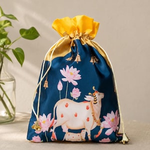 Puede incluir: Una bolsa con cordón de color azul oscuro que presenta un diseño estampado de una vaca blanca con huellas de manos rojas, un ternero y flores de loto rosas. La bolsa tiene una parte superior amarilla brillante, cordones dorados y pequeños dijes.