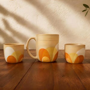 Peut inclure: Un mug en céramique avec une anse et deux petites tasses assorties, présentant une base couleur crème avec un motif peint de formes semi-circulaires orange, jaunes et blanches superposées, disposées en arc sur la partie inférieure.