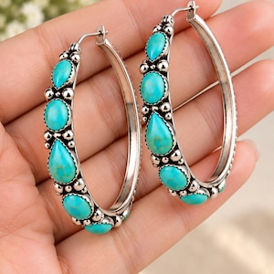 May include: A pair of silver hoop earrings featuring inlaid turquoise stones in various shapes, including teardrops and circles. The silver metalwork includes small decorative spheres between each stone, creating a textured border along the front of the hoops.