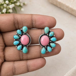 May include: A pair of stud earrings featuring a central oval pink stone surrounded by a fan-shaped arrangement of small, teardrop-shaped turquoise stones set in silver metal with a twisted rope border and beaded detailing around the edges.