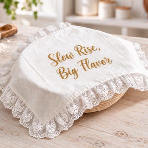 May include: A rectangular off-white linen bread proofing cloth featuring a ruffled white lace border with a heart pattern. The center of the cloth is embroidered with the text Slow Rise, Big Flavor in a golden-brown cursive script font.