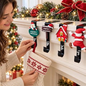 May include: Four black metal stocking holders with decorative Christmas-themed toppers including Santa, a Merry Christmas sign, a house, and a jumping Santa. Each holder features a vertical name tag with the names Zoe, Ella, Jude, and Ezra.