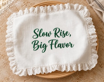 Puede incluir: Un paño rectangular blanco con un borde de encaje de ojales fruncido. El centro presenta un texto bordado en verde oscuro que dice "Slow Rise, Big Flavor" en una fuente cursiva. La tela tiene un tejido visible y un borde acabado.