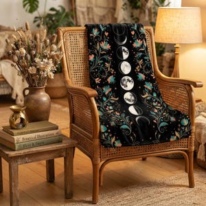 May include: A black throw blanket featuring a vertical column of moon phases in white and gray, surrounded by a colorful floral pattern with teal, pink, and orange blossoms and swirling vines, draped over a light brown woven wooden armchair.