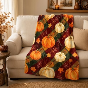 May include: A soft throw blanket featuring a repeating pattern of orange and cream-colored pumpkins set against a dark red and green plaid background, accented with small scattered autumn leaves and thin brown twig illustrations throughout the fabric.