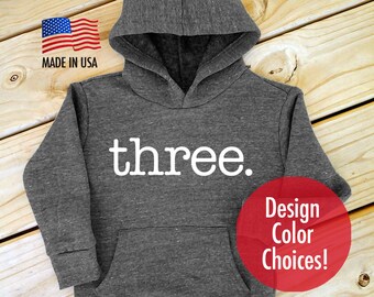 May include: Gray hooded sweatshirt with the word "three" printed in white on the front. The sweatshirt has a pocket on the front and is made in the USA. The text "Design Color Choices!" is printed in red on a white circle.