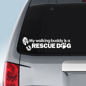 Puede incluir: Calcomanía de vinilo blanco para la ventana de un coche. La calcomanía dice "My walking buddy is a RESCUE DOG" con una huella de pata y huellas.