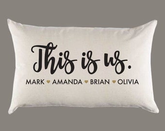 May include: A white pillow with the text "This is us." in black cursive lettering. Below the text are the names "Mark", "Amanda", "Brian", and "Olivia" with a heart symbol between each name.