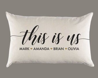 May include: A white pillow with the text "This is us" in black cursive lettering. Below the text are the names "Mark", "Amanda", "Brian", and "Olivia" with a gold heart symbol between each name.