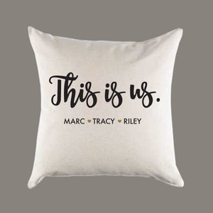 May include: A white throw pillow with the text "This is us." and the names "Marc, Tracy, Riley" printed in black with a gold heart symbol between each name.