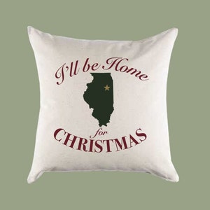 Może przedstawiać: Biała poduszka z napisem "I'll be home for Christmas" w czerwonych literach. Tekst jest wyśrodkowany na poduszce. Zielony zarys stanu Illinois jest wyśrodkowany na poduszce ze złotą gwiazdą w środku stanu.