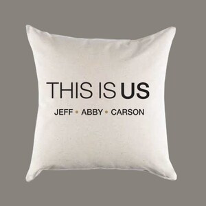 Op de afbeelding: Wit kussen met zwarte tekst die "This is Us" en "Jeff, Abby, Carson" zegt.