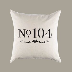 Op de afbeelding: Een wit kussen met zwarte tekst die "No. 104" luidt met een decoratieve krul eronder.