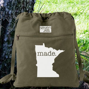 May include: A green canvas drawstring backpack with a white outline of the state of Minnesota and the text "made."  The backpack has a zippered pocket on the front with a white label that reads "seven miles per second".