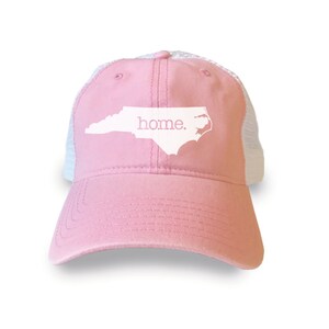 May include: Pink and white mesh trucker hat with a white outline of the state of North Carolina and the word "home." printed on the front.
