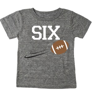 May include: Gray short-sleeved t-shirt with the word "SIX" in white letters and a brown football graphic. The football is being thrown with a white line behind it.
