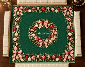 Puede incluir: Un tapete de mahjong verde cuadrado con una corona navideña central, lazos rosas, adornos rojos y dorados, y el texto The Millers. El borde está decorado con adornos y lazos a juego, rodeado por cuatro soportes para fichas blancos.