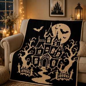May include: A black and cream knit throw blanket featuring a large haunted house design, a full moon, bare trees, and flying bats. The blanket has a solid cream-colored border and is draped over a sofa, displaying a detailed graphic pattern.