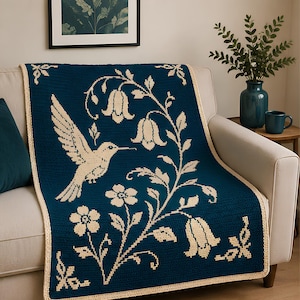 Puede incluir: Una manta de ganchillo de color verde azulado oscuro con un colibrí de color crema en vuelo junto a una enredadera con flores en forma de campana y pequeñas flores. La manta rectangular tiene un borde sencillo de color crema.