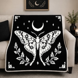 May include: A black throw blanket featuring a white pixelated moth graphic centred below a crescent moon. The design includes small stars and leafy branches on both sides, with decorative corner accents and a white border trim.