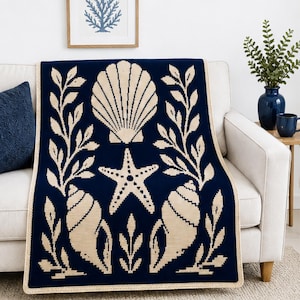 May include: A rectangular knit throw blanket featuring a deep navy blue background with a cream-colored pattern of a large central scallop shell, a starfish, two spiral seashells, and leafy botanical branches along the vertical edges.
