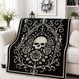 Pode incluir: Uma manta tecida em preto e creme com um motivo central de caveira rodeado por rosas detalhadas e padrões de arco em estilo gótico ornamentado. O design é simétrico com vinhas florais complexas e uma moldura de borda sólida.