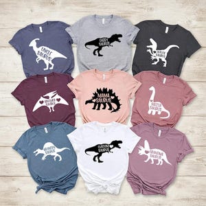 May include: A collection of nine short-sleeve t-shirts in various colors, each featuring a white or black dinosaur silhouette graphic. The prints include text such as Uncle Saurus, Daddy Saurus, Auntie Saurus, Brother Saurus, Mama Saurus, Sister Saurus, Grandpa Saurus, Custom Saurus, and Grandma Saurus.