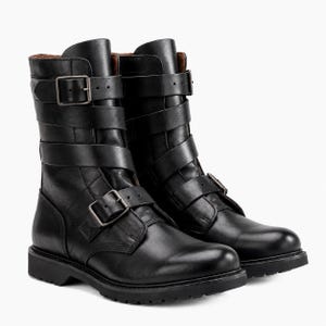 Puede incluir: Un par de botas negras de media caña con acabado en cuero liso, puntera redondeada y suela de goma gruesa con tacos. Cada bota se asegura con tres correas horizontales en la caña y una correa sobre el empeine, todas con hebillas metálicas.