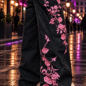 Könnte beinhalten: Schwarze Jeans aus Denim mit weitem Bein und einem auffälligen, rosafarbenen Blumenstickmuster, das sich an der Seite des Beins hochzieht. Die Hose verfügt über einen Standardbund mit Gürtelschlaufen und ein klassisches Fünf-Taschen-Design.