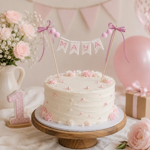 Può includere: Una torta rotonda con glassa bianca decorata con piccoli fiori di zucchero rosa e bianchi e perle. Un topper per torta presenta quattro bandierine in tessuto bianco che compongono la scritta MAYA in filo rosa, sospese tra due bastoncini con fiocchi.