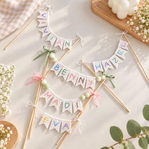 May include: Four cake toppers featuring white felt pennant flags strung on wooden sticks. Each banner displays embroidered names including VIVIAN, BENNY, HAZEL, MADI, and EVA in colourful thread. The designs are accented with small satin ribbons and beads.