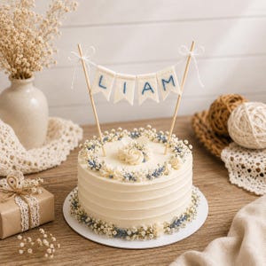 May include: A round white frosted cake decorated with small white and blue flowers, topped with a felt banner featuring the name LIAM in blue stitching. The banner is suspended between two wooden sticks with white bows at the top.