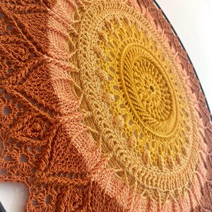 Op de afbeelding: Een ronde gehaakte wandhanger met een ingewikkeld mandalapatroon. Het ontwerp verloopt van een felgeel centrum naar tinten oranje en dieproest aan de buitenranden, allemaal gemonteerd in een dunne, zwarte, ronde omlijsting.
