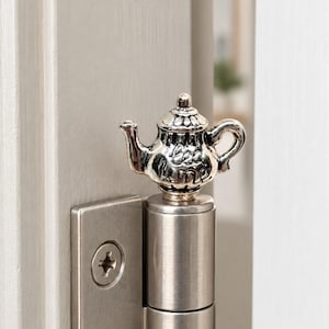 Peut inclure: Un embout de charnière de porte en métal argenté en forme de théière miniature. La théière présente un bec, une anse, un couvercle détaillés et le texte en relief "tea time" sur le côté. L'embout est fixé au sommet d'une charnière en métal brossé.