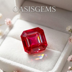 Puede incluir: Una gema roja de corte cuadrado con superficies facetadas que reflejan la luz, exhibida dentro de un joyero forrado en terciopelo blanco. El texto OASISGEMS es visible en la parte superior. Pequeñas flores blancas rodean el joyero abierto.