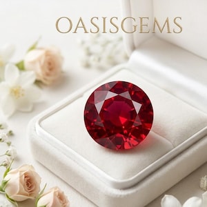 Pode incluir: Uma pedra preciosa redonda facetada de cor vermelho profundo está dentro de uma caixa de joias de veludo branco. O texto OASISGEMS está impresso em dourado acima da caixa. Pequenas flores brancas e rosa pálido rodeiam a caixa aberta.