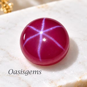 Puede incluir: Una piedra preciosa de rubí estrella de corte cabujón redondo, de color magenta intenso, con un efecto de estrella blanca de seis rayos centrado en su superficie. El texto Oasisgems está impreso en negro sobre el mármol blanco bajo la piedra.