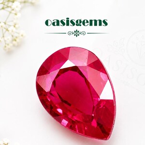 Könnte beinhalten: Ein birnenförmiger, facettierter Edelstein in einer leuchtenden, tiefroten Farbe. Der Stein weist einen Brillantschliff mit mehreren reflektierenden Oberflächen auf. Der Text oasisgems ist in Dunkelgrün über dem Stein abgedruckt.