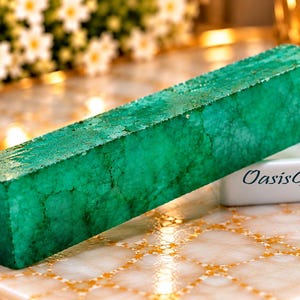 May include: A rectangular emerald green gemstone block with a textured, crystalline surface. The stone features deep green hues and natural internal fractures. A small white rectangular label with the black text OasisGems is visible.