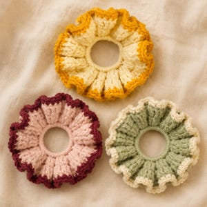 May include: Three circular crochet hair scrunchies with ruffled edges. One is yellow and cream, one is dusty pink with a dark maroon border, and one is sage green with a cream border. Each features a textured, hand-stitched yarn construction.