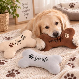 May include: Three plush bone-shaped dog toys in beige, brown, and light gray. Each toy features a dark brown paw print icon and a personalized name embroidered in black script: Capello on the beige toy, Antonio on the brown, and Daniela on the gray.