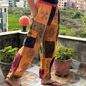 May include: Loose-fitting pants featuring a patchwork design in shades of orange, brown, and maroon. The fabric displays various black printed symbols and patterns, including a side cargo pocket, an elastic waistband, and a relaxed silhouette.