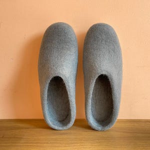 以下が含まれることがあります： 丸みを帯びた爪先とスリッポン型のデザインが特徴の、ライトグレーのフェルトウール製スリッパ一足。スリッパは質感のあるマットな表面をしており、外側から内側まで一貫したニュートラルなグレーの色合いで構成されています。