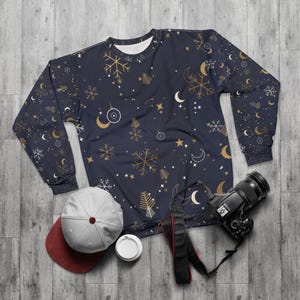 May include: A navy blue long-sleeve sweatshirt featuring a repeating pattern of gold and white snowflakes, crescent moons, stars, and circular ornaments. A light gray baseball cap with a maroon brim and a black digital camera are positioned below.