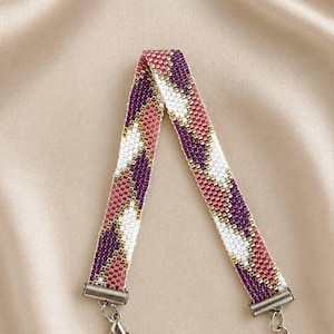 May include: A woven beaded bracelet featuring a geometric pattern of diagonal stripes in shades of deep purple, dusty rose, and white. The band is secured with silver-toned metal end caps, a lobster claw clasp, and an adjustable chain extension.