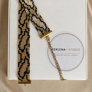 May include: A beaded choker necklace featuring a repeating snake-skin pattern in black, tan, and cream glass beads. The band is secured with gold-toned metal end bars and includes a gold-toned chain extender for adjustable length and a secure clasp.